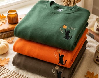 May include: A stack of three crewneck sweatshirts in dark green, orange, and dark brown. Each sweatshirt features an embroidered design of a black cat reaching up toward a falling autumn leaf with swirling lines, positioned on the upper chest area.