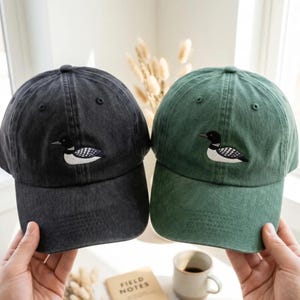 Puede incluir: Dos gorras de béisbol, una en gris carbón y otra en verde bosque, cada una con un colimbo bordado en el panel frontal. Las gorras tienen un acabado de tela lavada y texturizada, una visera curva y una corona estructurada con ojales de ventilación.