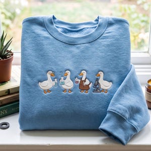 Peut inclure: Un sweat-shirt bleu clair à col rond orné d'une broderie représentant quatre canards blancs effectuant des expériences scientifiques. Les canards sont illustrés avec du matériel de laboratoire, notamment des tubes à essai, des béchers et un microscope.