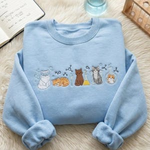 Könnte beinhalten: Ein hellblaues Sweatshirt mit Rundhalsausschnitt und einer Stickerei, die verschiedene Katzen bei der Interaktion mit Laborgeräten wie Bechern, Reagenzgläsern und chemischen Formeln wie H2O und O2 zeigt, auf weichem Stoff.