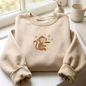 May include: A beige crewneck sweatshirt featuring an embroidered graphic of a brown monkey holding a white mug, surrounded by small gold stars. The sweatshirt has long sleeves with ribbed cuffs and a ribbed neckline, crafted from a soft fabric.