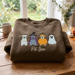 May include: A brown crewneck sweatshirt featuring an embroidered design of four dogs in Halloween costumes: a mummy, a witch, a pumpkin, and a ghost. Below the dogs, the text Pit Boo is embroidered in a white cursive script font.