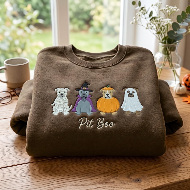 May include: A brown crewneck sweatshirt featuring an embroidered design of four dogs in Halloween costumes: a mummy, a witch, a pumpkin, and a ghost. Below the dogs, the text Pit Boo is embroidered in a white cursive script font.