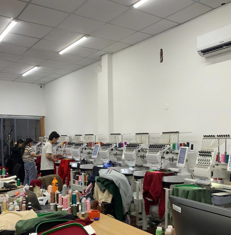 May include: A commercial embroidery workshop featuring a row of white industrial embroidery machines with multiple needles and digital screens. Various colorful thread spools are arranged on tables, and garments are positioned under the needles.