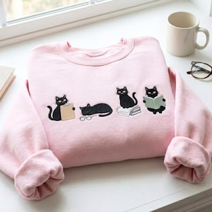 Puede incluir: Una sudadera de cuello redondo en color rosa claro con cuatro gatos negros bordados. Un gato sostiene un portapapeles, otro duerme junto a unas gafas, otro está sentado sobre una pila de papeles y el último lee un libro. El diseño está centrado.
