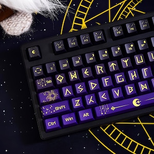 May include: A mechanical keyboard featuring dark purple keycaps with yellow celestial and runic symbols. The keys display moon phases, stars, and geometric patterns. The spacebar shows a crescent moon and star design, while the function keys include moon icons. The layout includes standard alphanumeric keys alongside specialized mystical iconography.