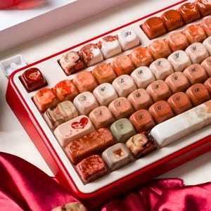 May include: A mechanical keyboard featuring custom keycaps in shades of red, orange, and cream. Several keys display floral patterns, marble textures, and a heart graphic. One key is printed with the text 'Passionate as fire' in a cursive font.