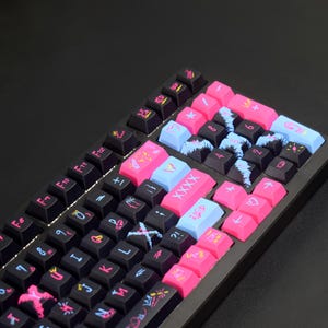 Op de afbeelding: Een mechanisch toetsenbord met zwarte toetsen voorzien van kleurrijke grafische prints in roze, blauw en geel. De toetsen tonen letters, cijfers en symbolen, waaronder hartjes, kronen en de tekst XXXX.