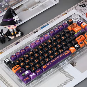 May include: A mechanical keyboard with a transparent frame and custom keycaps featuring a Halloween theme. The keys are black, purple, and orange, displaying letters, numbers, and icons like pumpkins, ghosts, and bones. The spacebar reads ALL SAINTS' DAY.