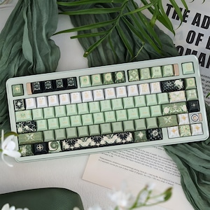 May include: A mechanical keyboard with a light green frame and a mix of light green, white, and black keycaps. The keys feature various botanical, celestial, and floral illustrations, including stars, moons, butterflies, and roses, with standard QWERTY layout.