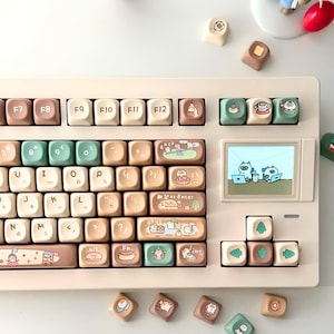 May include: A mechanical keyboard with a cream-colored frame and custom keycaps in shades of beige, brown, and sage green. The keys feature cartoon illustrations of small animals, food items, and a small digital screen displaying two characters.