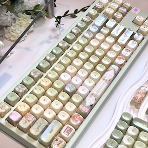 May include: A mechanical keyboard featuring a sage green frame and a set of rounded keycaps in shades of cream, sage green, and olive. Several keycaps display printed illustrations of small figures and floral patterns, with a long spacebar.