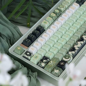 May include: A mechanical keyboard featuring a sage green frame and a custom keycap set. The keys display a mix of light green, white, and black colors with floral patterns, celestial star motifs, moon phases, and standard alphanumeric characters.