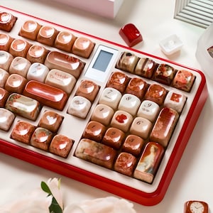 Puede incluir: Un teclado mecánico con marco rojo y una mezcla de teclas brillantes, color crema y marrón. Algunas teclas presentan motivos florales, corazones y el retrato de una persona. Una pequeña pantalla digital está integrada en la parte superior central.