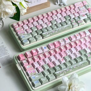 Puede incluir: Dos teclados mecánicos con marcos de color verde salvia y teclas personalizadas. Las teclas presentan una paleta de colores rosa suave y verde salvia, decoradas con diversas ilustraciones florales, botánicas y de nubes, con letras QWERTY.