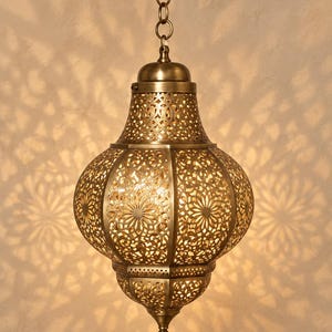 May include: A brass-toned metal pendant light fixture with an intricate laser-cut floral pattern. The bulbous, onion-shaped lantern is suspended by a matching metal chain and canopy, casting detailed, symmetrical shadow patterns on the wall behind.
