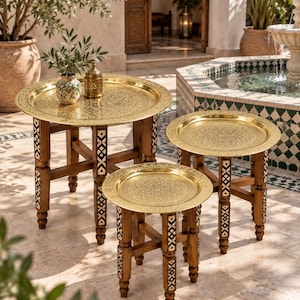 May include: A set of three round nesting tables with polished brass trays featuring intricate etched patterns. The wooden legs are inlaid with geometric mother-of-pearl accents. The tables vary in height and diameter, each supported by four sturdy legs connected by a cross-brace structure.