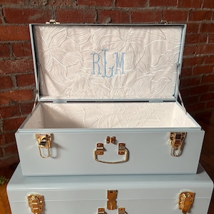 May include: A set of two light blue storage trunks with gold-toned metal latches and handles. The interior of the open top trunk is lined with white fabric featuring a floral pattern and the embroidered monogram letters RLM in light blue thread.