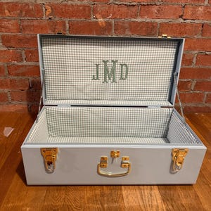 May include: A light blue storage trunk with gold-toned metal latches and a central handle. The interior is lined with a white and light blue gingham fabric, featuring the embroidered monogram letters JMD in a sage green thread.