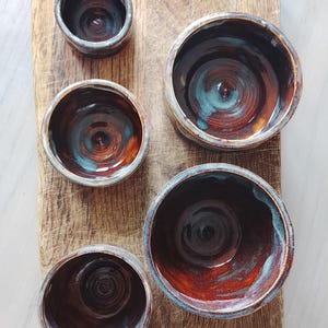 Könnte beinhalten: Ein Set aus fünf handgefertigten Keramikschalen in verschiedenen Größen mit einer glänzenden Glasur in tiefen Braun-, Kupfer- und Türkistönen. Die Schalen haben eine kreisförmige Form mit sichtbaren konzentrischen Ringen im Inneren.