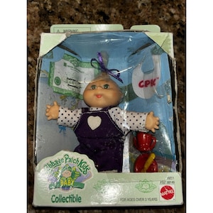 May include: A Cabbage Patch Kids collectible baby doll in original packaging. The doll has a soft face, green eyes, and wears a white long-sleeved shirt with black polka dots under purple overalls with a white heart. Includes a birth certificate.