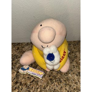 May include: A plush Ziggy doll with a large round head, a prominent nose, and a small black mouth. The doll wears a yellow shirt with the word Ziggy printed in red on the side. It holds a small white plush animal with a blue nose and black eyes.