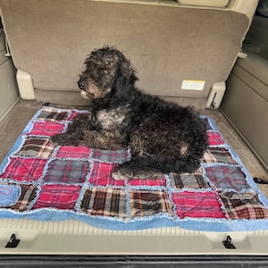 May include: A black curly-haired dog resting on a rectangular rag quilt. The quilt is constructed from square patches of red plaid, brown plaid, and blue denim fabric with frayed edges. The quilt is laid flat on the floor of a vehicle trunk.
