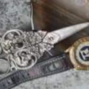 May include: A pair of silver-toned scissors with ornate, dragon-shaped handles and a gold-toned wristwatch featuring a black face, a gold bezel, and a dark, textured leather strap with decorative stitching and metallic accents.