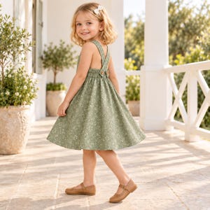 Pu&ograve; includere: Un vestito scamiciato verde salvia con un delicato motivo floreale bianco, vita elasticizzata arricciata e spalline incrociate con bottoni marroni, abbinato a scarpe Mary Jane in pelle color cuoio e una piccola molletta bianca.