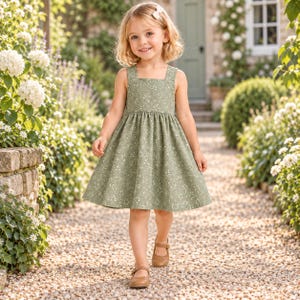 Pu&ograve; includere: Un abito senza maniche color verde salvia con scollo quadrato e un delicato motivo floreale bianco. L'abito presenta un corpetto aderente e una gonna arricciata e svasata che cade sopra il ginocchio. Abbinato a semplici scarpe in pelle marrone.