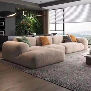 May include: A large, low-profile sectional sofa upholstered in a light beige fabric. The sofa features rounded edges and a modular design, accented with one green, one mustard yellow, and one dark gray throw pillow placed along the back cushions.