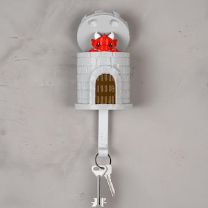May include: A gray wall-mounted key holder shaped like a stone tower with a brown arched door. A small red dragon with white horns peeks out from the top of the tower. A white hook extends from the bottom, holding two silver metal keys.