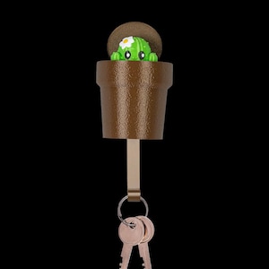 May include: A brown textured wall-mounted key holder shaped like a flower pot. A small green cactus with a white flower and black eyes peeks out from the top. A metal strip extends downward, holding a silver ring with two metallic keys.