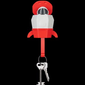 May include: A red and white rocket-shaped key holder featuring a small astronaut figure in the cockpit. The rocket has a red base with two side fins and a red top section. A red strap hangs from the bottom, holding a metal ring with two silver keys.