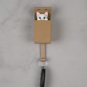 May include: A tan wall-mounted key holder featuring a small white cat figure with pink ears peeking out from the top compartment. A vertical tab extends downward from the base of the holder, supporting a silver metal ring holding a black car key fob.