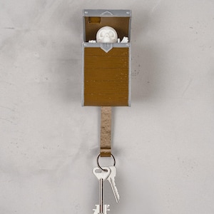 Puede incluir: Un llavero de pared con forma de cofre de madera marrón con borde plateado. Una figura de calavera blanca es visible dentro del cofre abierto. Una correa marrón texturizada cuelga de la parte inferior, sosteniendo dos llaves de metal plateado.
