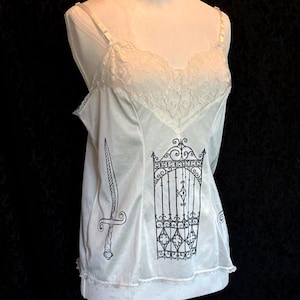 May include: A white camisole featuring a lace-trimmed neckline and thin adjustable straps. The front of the garment displays a black graphic print of an ornate iron gate in the center, flanked by a vertical sword illustration on the right side.