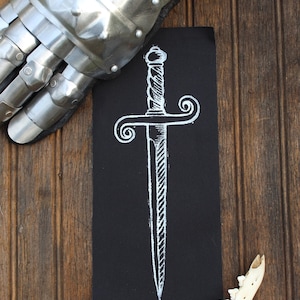May include: A rectangular black fabric patch featuring a white screen-printed illustration of a double-edged dagger with a spiraled handle and curved crossguard. The patch is displayed alongside a metallic gauntlet and a white animal jawbone.
