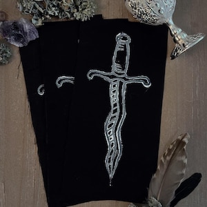 May include: Three rectangular black fabric patches, each featuring a hand-drawn white ink illustration of a wavy-bladed dagger. The patches are arranged in a slight overlap, showcasing the detailed line work of the silver-toned dagger design.