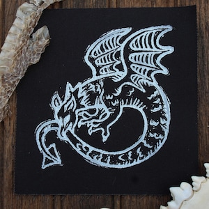 May include: A square black fabric patch featuring a white screen-printed graphic of a coiled dragon with wings and a pointed tail. The dragon is depicted in a stylized, linear art style with visible texture in the white ink lines.