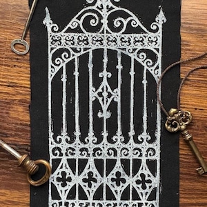 May include: A rectangular black fabric patch featuring a white screen-printed illustration of an ornate wrought iron gate with intricate scrollwork and pointed finials, accompanied by three antique-style metal keys in silver and brass tones.