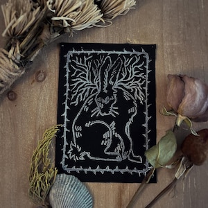 May include: A rectangular black fabric patch featuring a silver ink illustration of a rabbit with long, branch-like antlers. The rabbit is enclosed within a rectangular border composed of sharp, thorn-like spikes, creating a stark contrast.