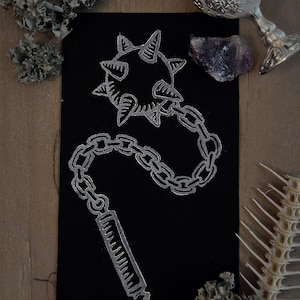 May include: A rectangular black fabric patch featuring a silver-colored embroidered design of a flail. The design depicts a spiked ball attached to a chain connected to a cylindrical handle, rendered in a metallic silver thread against the black fabric.