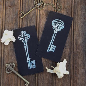 May include: Two rectangular black fabric patches featuring light blue screen-printed illustrations of ornate skeleton keys. The patches are displayed alongside two antique-style brass keys and two small, off-white, irregular bone-like objects.