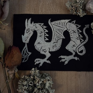 May include: A rectangular black fabric patch featuring a silver-colored screen-printed graphic of a dragon in profile. The dragon is depicted with wings, a long tail, and flames emerging from its mouth, rendered in a stylized, linear design.
