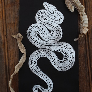 May include: A rectangular black fabric patch featuring a white screen-printed illustration of a coiled snake with detailed scales. The patch is surrounded by two pieces of translucent, textured, shed snake skin on a dark wooden surface.