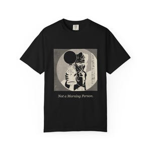 May include: A black short-sleeve t-shirt featuring a graphic print of a stylized figure holding a perfume atomizer within a circular frame. Below the graphic, the text reads, Not a Morning Person. The design uses shades of black, white, and gray.