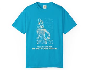 May include: A bright blue short-sleeve t-shirt featuring a white line-art illustration of a puppet character with a long nose. Below the graphic, the text reads: PULL MY STRINGS. SEE WHAT F*CKING HAPPENS. The shirt has a crew neckline.