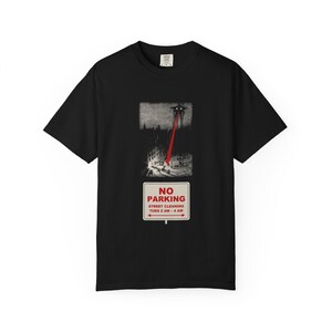 Pode incluir: Uma t-shirt preta de manga curta com um gráfico de uma máquina tripé disparando um raio laser vermelho em uma rua. Abaixo da ilustração há uma placa retangular branca que diz NO PARKING STREET CLEANING TUES 2 AM - 4 AM.