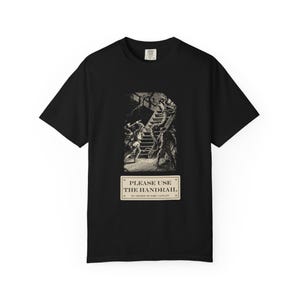 Puede incluir: Una camiseta negra de manga corta con una ilustración de estilo antiguo que muestra figuras en una escalera con tentáculos. Debajo del gráfico hay un letrero rectangular con el texto: PLEASE USE THE HANDRAIL BY ORDER OF THE CAPTAIN.